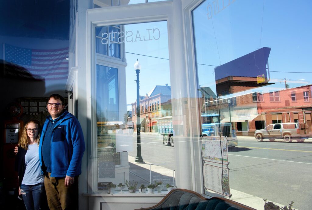 Two people standing inside a storefront with large windows, reflecting a quiet street scene in a small town.