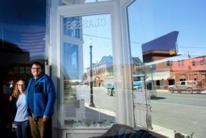 Two people standing inside a storefront with large windows, reflecting a quiet street scene in a small town.