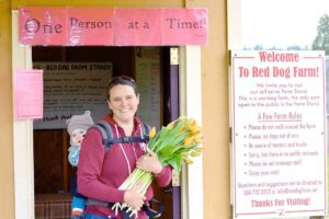 Smiling person holding a bouquet of tulips with a baby in a carrier, standing at the entrance of Red Dog Farm.