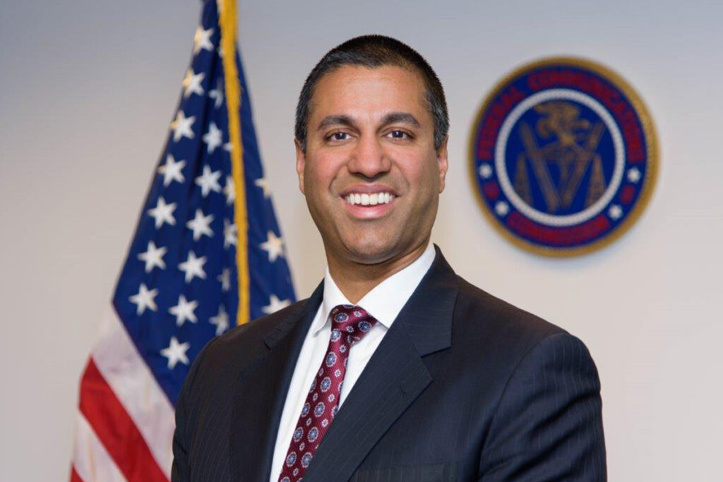 Person in a suit smiling in front of a U.S. flag and a government agency seal on the wall.