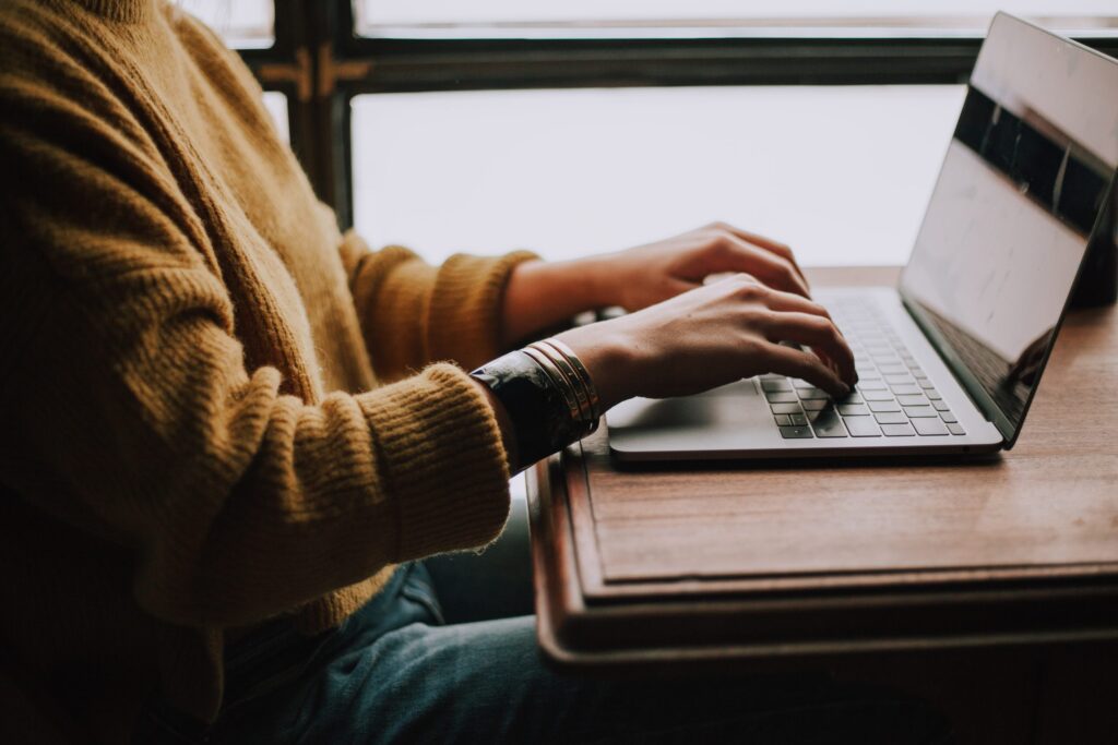 Person in a mustard sweater typing on a laptop at a wooden desk near a window.