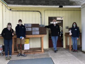 Four people wearing masks standing outside a building near boxes stacked on tables, possibly for a distribution or donation event.