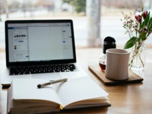Open notebook with a pen, laptop, coffee mug, and small flower vase on a wooden desk by a window.