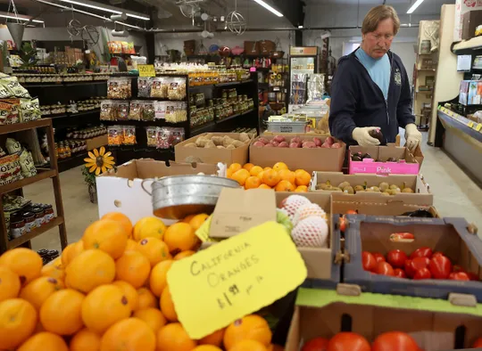 Person shopping in a grocery store, standing near boxes of fresh produce including oranges, tomatoes, kiwis, and potatoes.