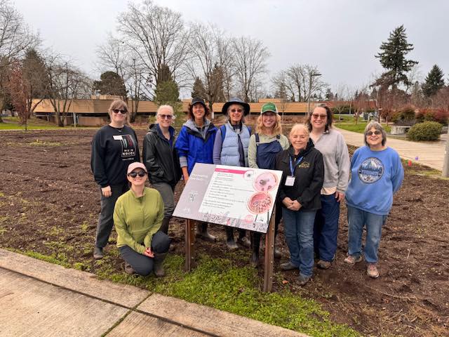 Group of Master Gardeners standing outdoors around an educational sign in a garden space