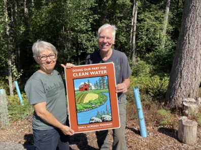 Two people holding a Doin Our Part for Clean Water sign with a sloped forest and seedling cages in the background