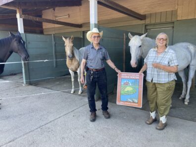 Two people in a horse shelter holding a Doing Our Part for Clean Water sign with three horses beside them.