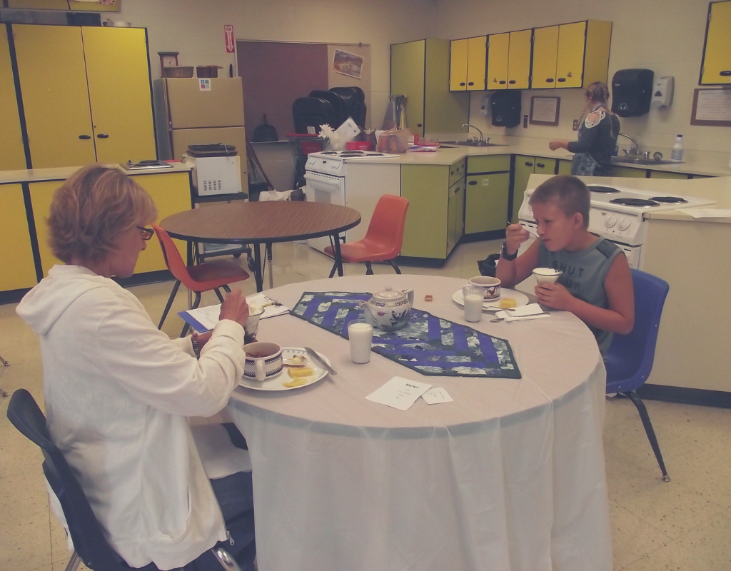 Adult and child sitting at a table eating in a classroom kitchen while another person works at the counter in the background.