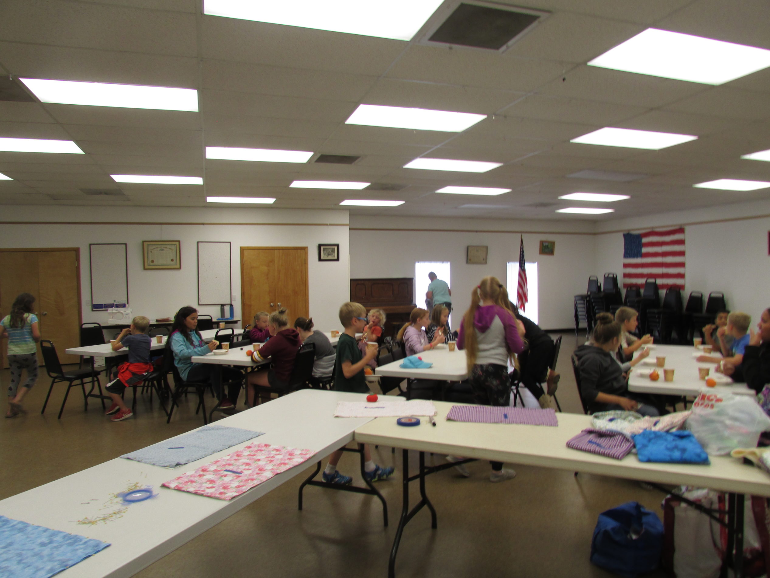 Group of children sitting at tables in a large community room, working on activities and eating snacks.