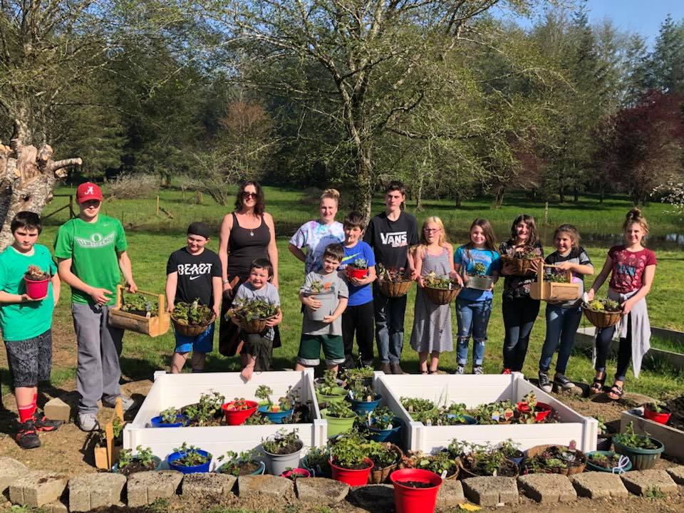 Group of youth and an adult standing outdoors in a garden, holding potted plants in front of raised garden beds on a sunny day.