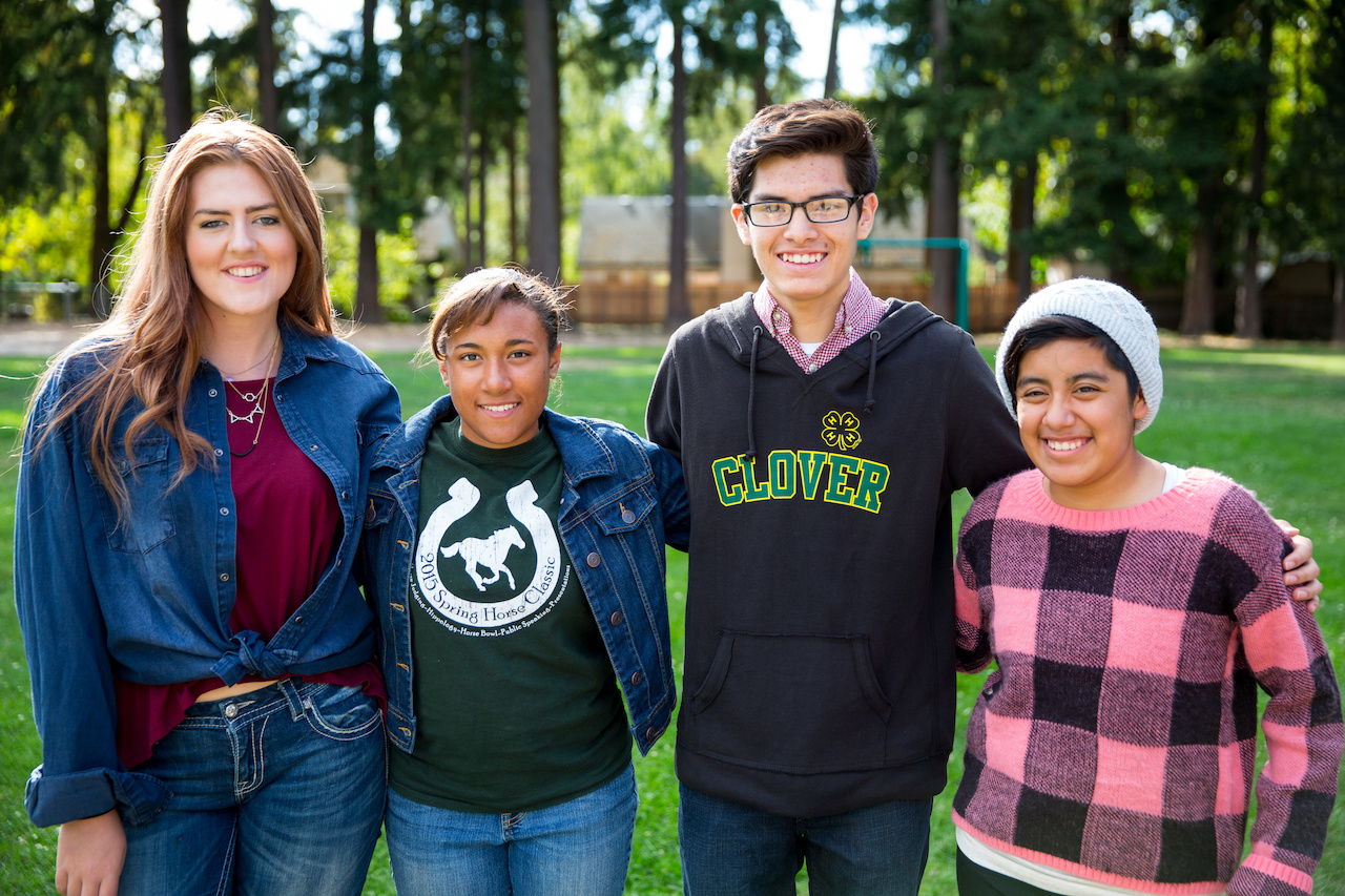 Four smiling teenagers standing together outdoors in a park, with trees in the background, wearing casual clothes including a 4-H hoodie.
