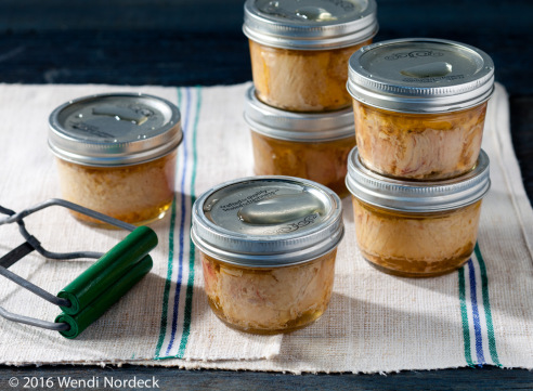 Several small glass jars with metal lids filled with preserved food, arranged on a cloth with canning tongs nearby.