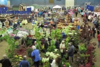 Large indoor plant and garden show with many people browsing tables filled with flowers, shrubs, and gardening displays.