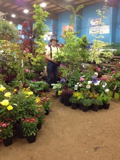 Person standing among rows of potted flowers and plants inside an indoor plant sale or nursery display.