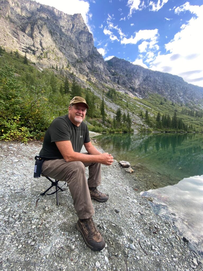 A man wearing a cap and hiking clothes sits on a small folding stool by a clear mountain lake, smiling at the camera, with steep rocky cliffs and pine trees rising behind him under a partly cloudy sky.