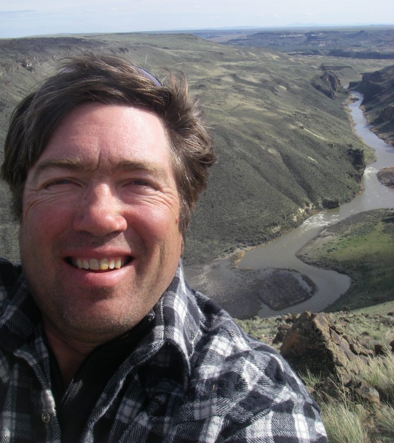 Steve Stuebner smiling while taking a selfie with a mountain landscape behind him, where a river winds through the valley below.