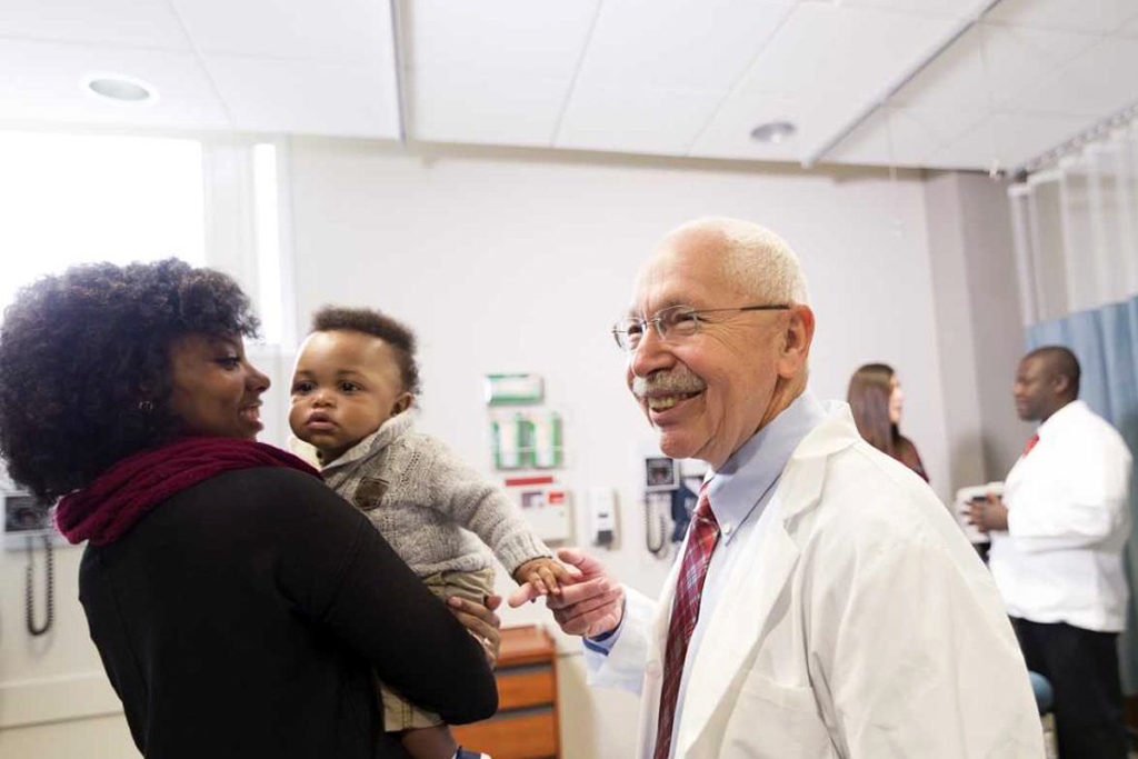 Left a mother is holding a baby in her arms while smiling. Right Dr. George Novan holds the hand of the baby while smiling.