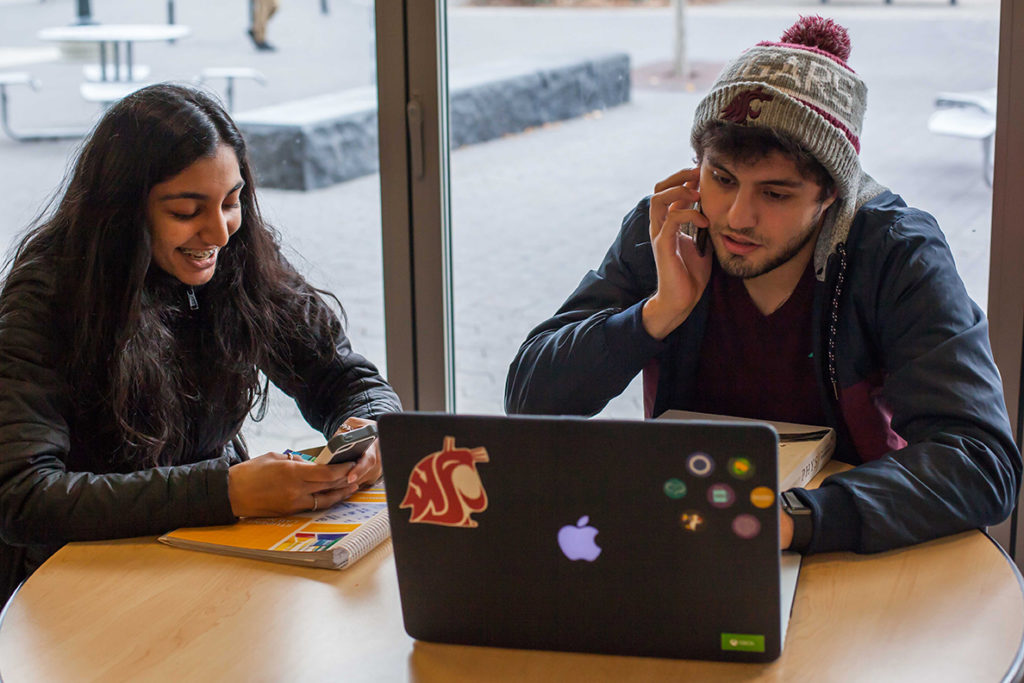 Two students sitting at table looking at a phone and a computer.