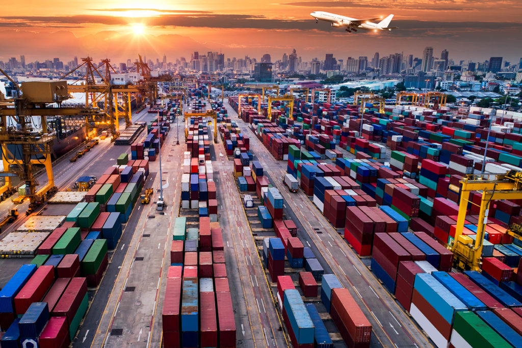 Cranes tend to cargo in a container yard as a plane flies overhead.