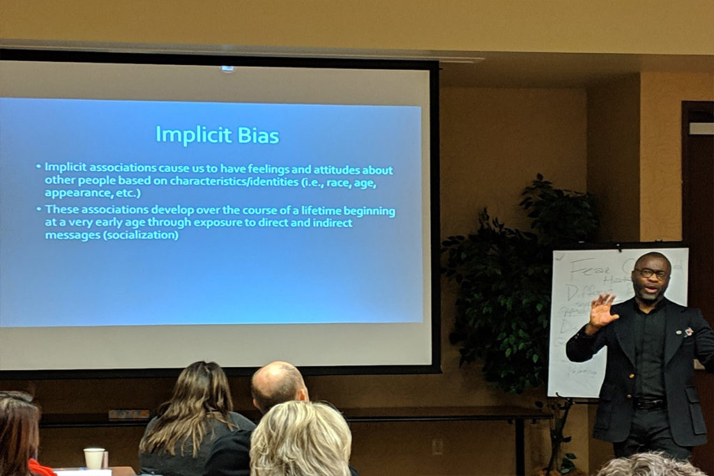 Obie Ford III discussing implicit bias with a slideshow presentation on a projector screen beside him.