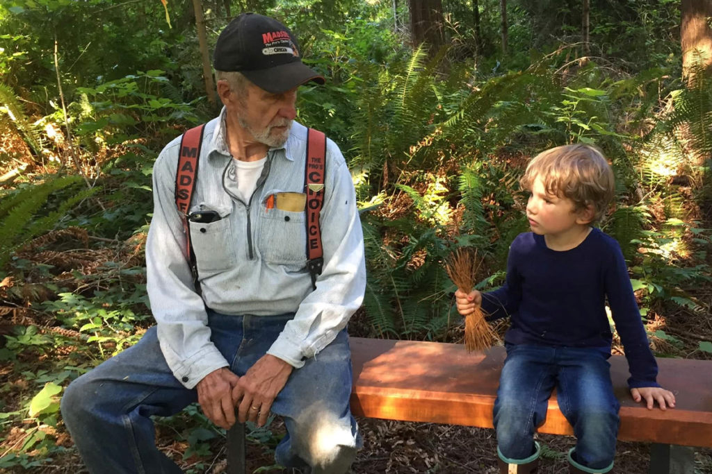 A man and a child sit on a log in a forest.