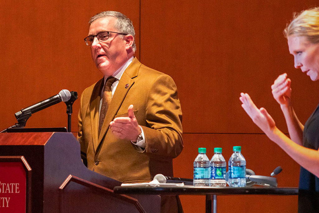Kirk Schulz speaking at a podium while a sign language interpreter stands nearby.