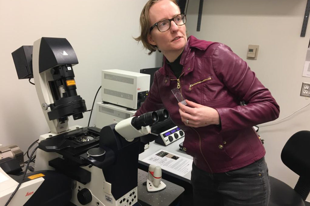 Beverley Rabbitts stands next to an electron microscope holding a specimen slide.