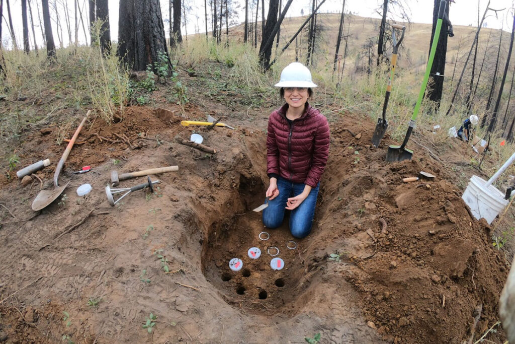 A woman in a hard hat kneeling before a soil pit dug on a burned forest landscape