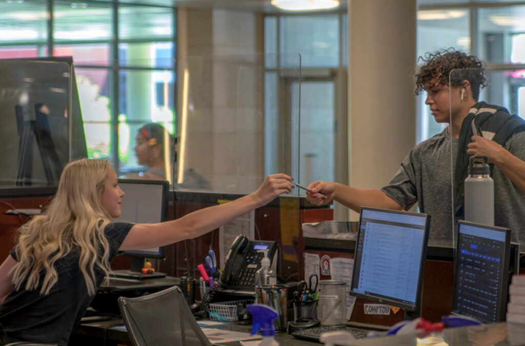 A student employee working at an information desk reaches for a pen from someone.
