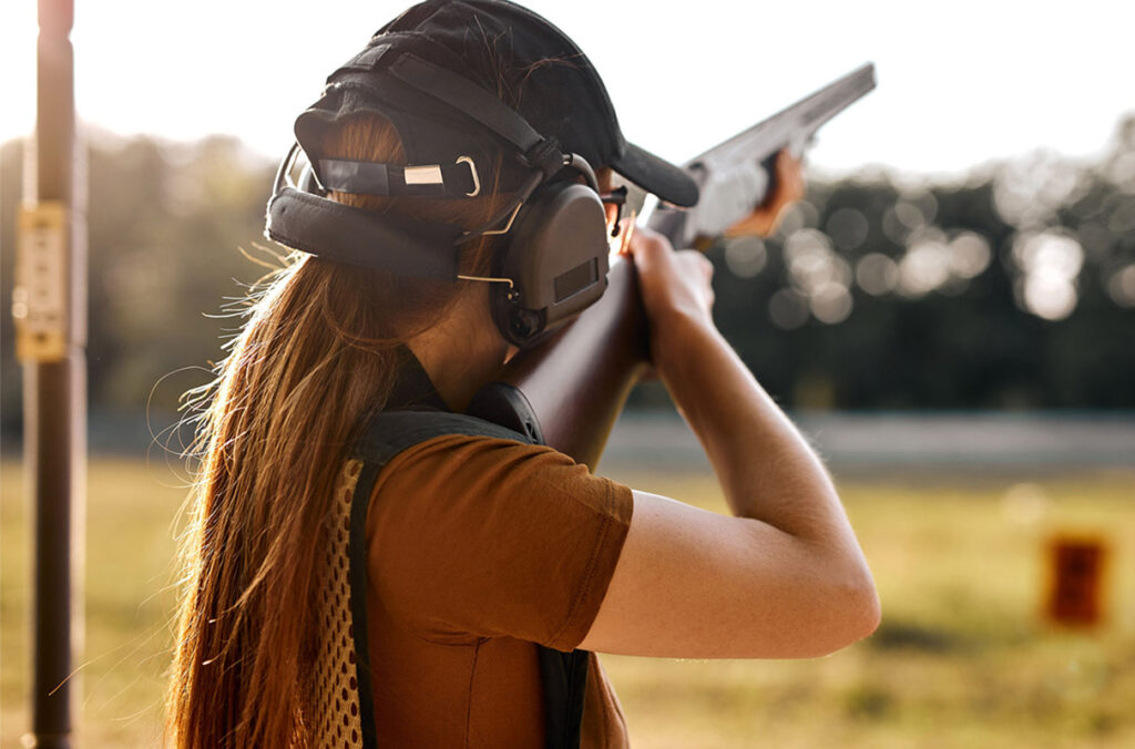 A teenage girl seen from behind prepares to fire a hunting rifle.