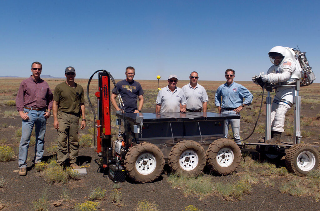 Engineers, astronauts, and civilian technicians posing next to equipment to be used on the moon and Mars.