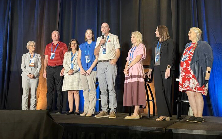 Eight people standing on a stage as one speaks into a microphone.