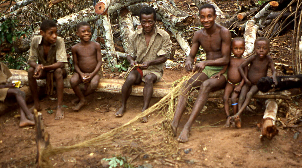 African men and children sitting on logs as a man demonstrates how to weave a hunting net.