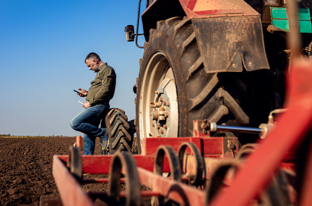 A farmer leaning against a tractor tire looking at his cell phone while holding a wrench.