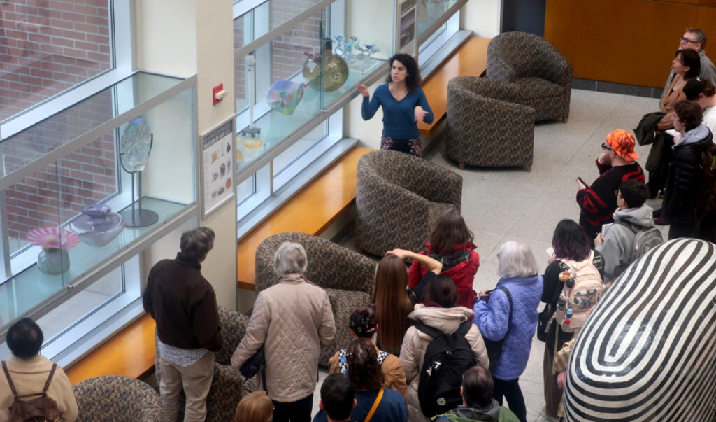 A small crowd listening to someone explaining various pieces of glass artwork.