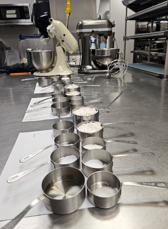 Rows of metal measuring cups with various ingredients sitting on a countertop.