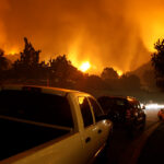 Two people sit on a curb amongst parked cars watching a nearby wildfire threaten their neighborhood.