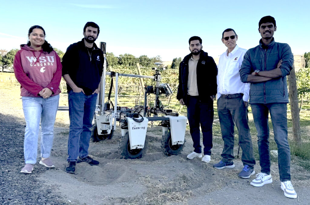 Five people standing next to an ag robot in front of an apple orchard.