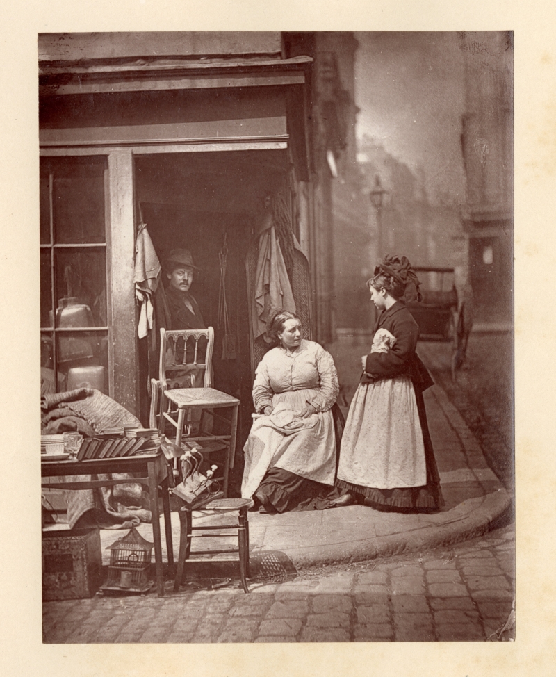 A Woodburytype print of a man in a doorway and two women, one of them seated, on a London street corner.