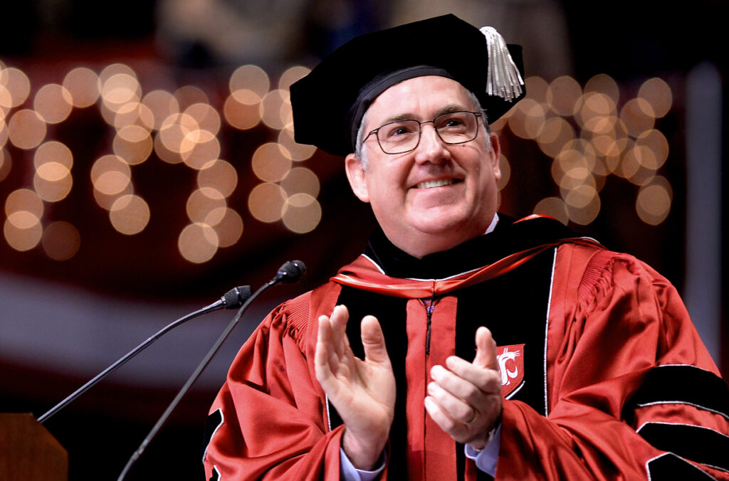 WSU President Kirk Schulz standing at a podium and clapping during a commencement ceremony.