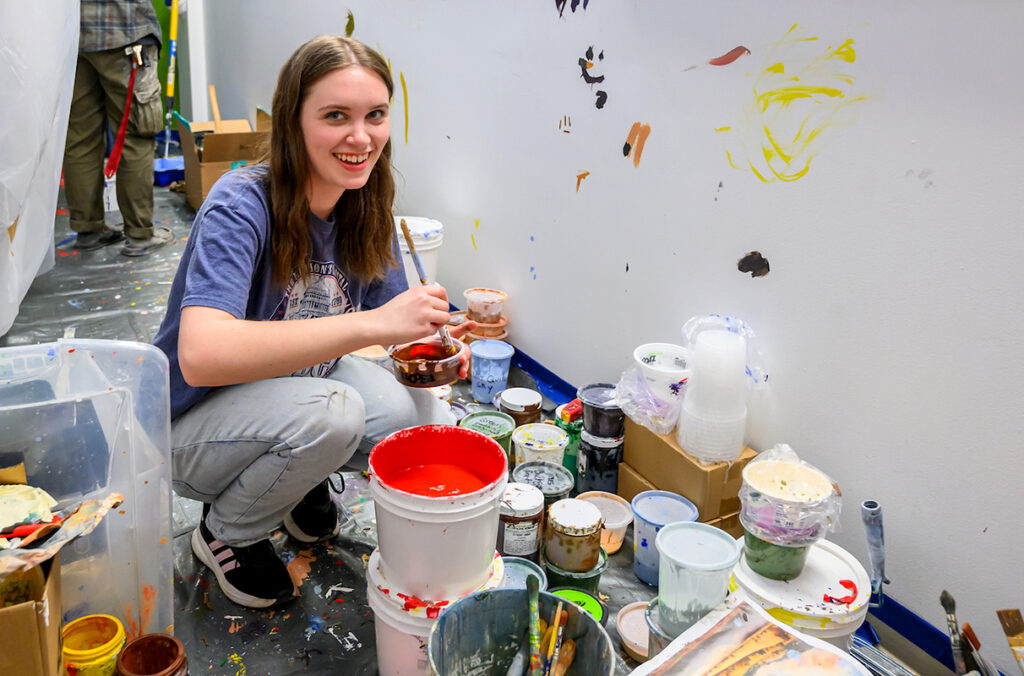 Mycala Zimmerman squatting next to a stack of art supplies as she mixes paint.