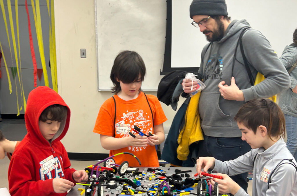 A parent watches as three children build with LEGO bricks.