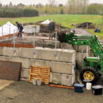 Three men standing on concrete walls as a fourth operates a tractor during construction of a new compost plant.