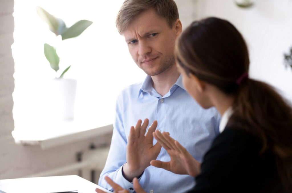 A male employee with a slight frown putting his hands up in front of him as he looks at a female coworker.