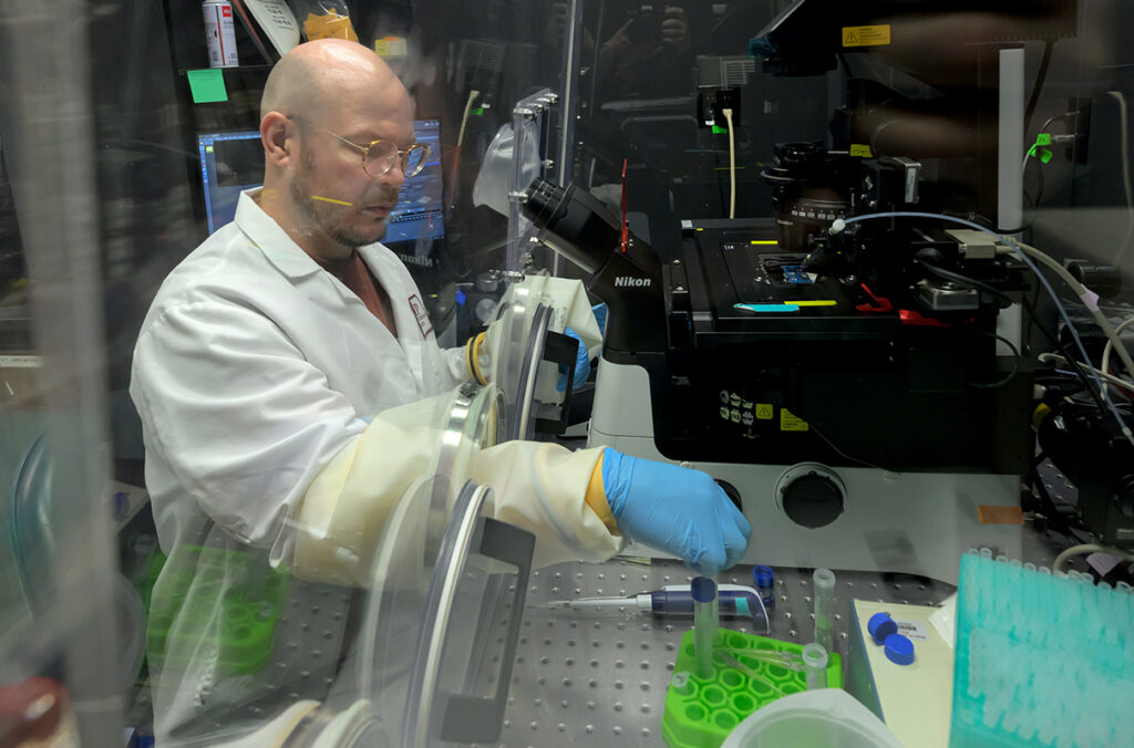 Arden Baylink reaching through a plastic barrier to use a microscope in a research lab.
