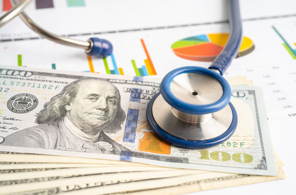 A stethoscope lies on top of $100 bills and pieces of paper displaying graphs.