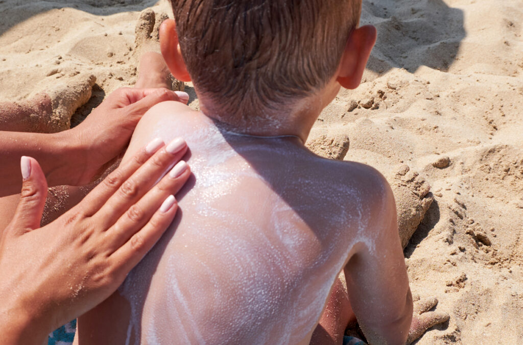 A mother's hand applying sunscreen to the back of her son at the beach.