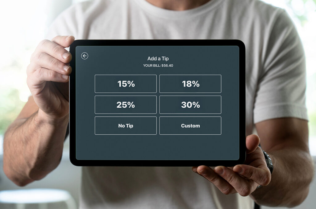 Closeup of a man holding a tablet showing a bill and various tipping options and amounts.