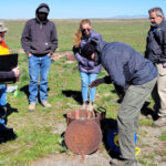 Five people watch as someone runs measuring tape down a monitoring well.