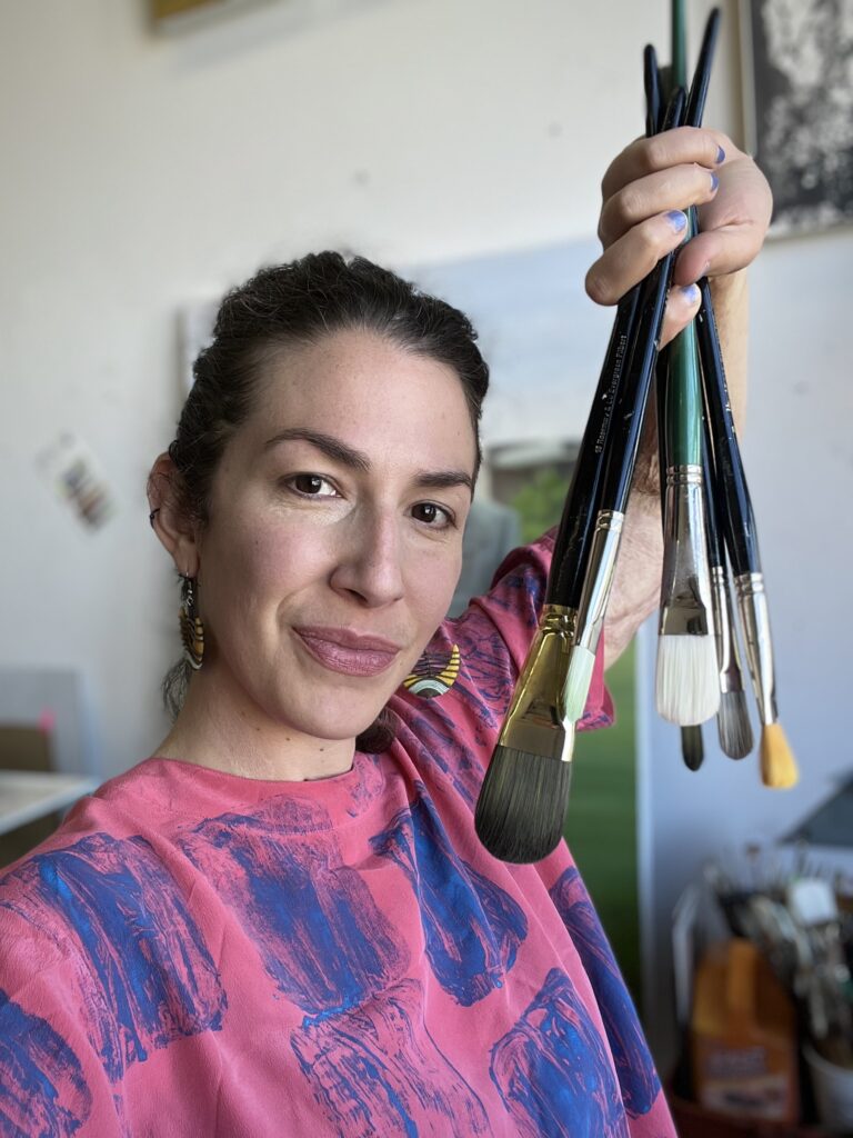 Artist Grace Flott posing with paint brushes in front of a portrait of WSU President Kirk Schulz.